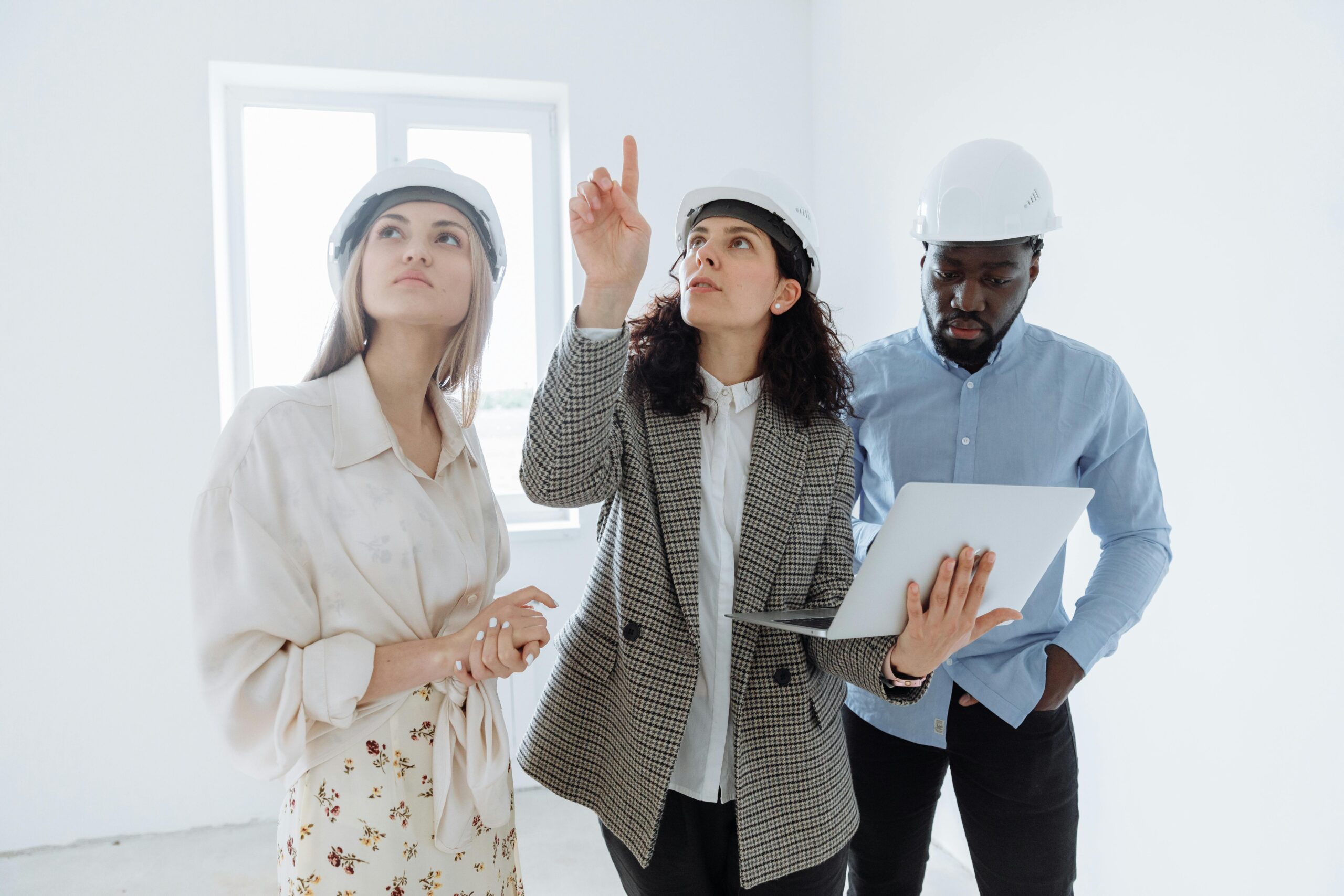 A diverse team of architects wearing helmets discusses interior plans with a laptop in a bright room.